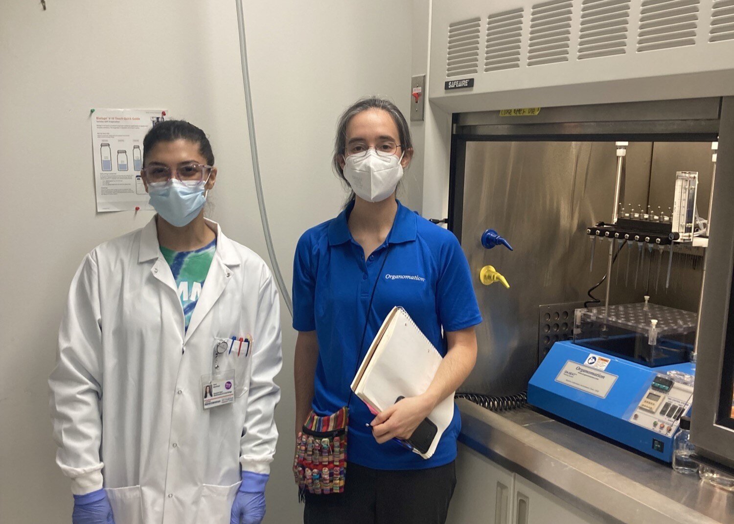 Two woman in a laboratory standing next to a fume hood with an Organomation MULTIVAP evaporator inside. One woman is wearing a lab coat, and another a blue Organomation polo. Both women are wearing masks.