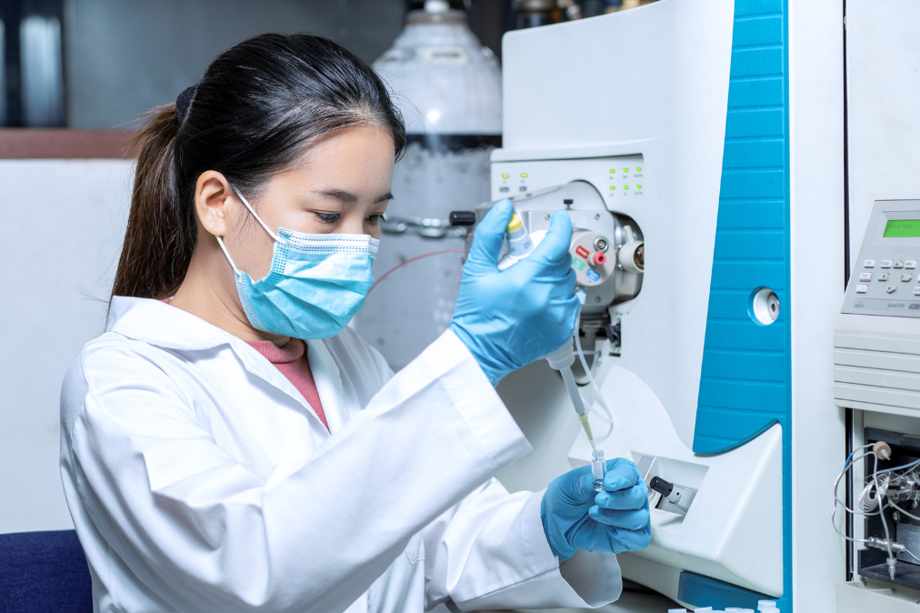 a young woman of Asian descent wearing a blue surgical mask and blue gloves as she uses a syringe to transfer some green liquid into a small vial