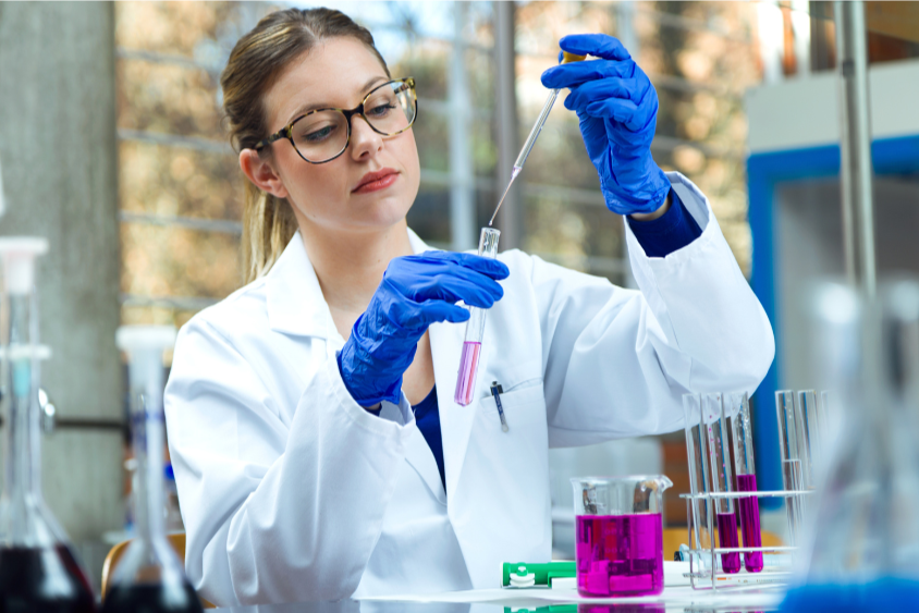 a white blonde woman wearing blue gloves and a white lab coat using a dropper to transfer a pink solvent from a beaker to a test tube