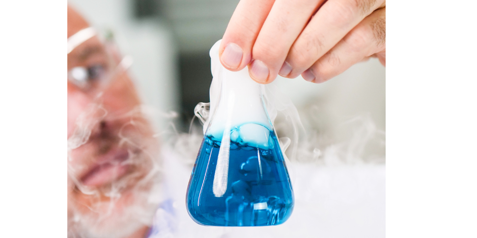 an older white man with a short gray beard holding up a beaker which is filled with blue solvent that is producing white bubbles and overflowing