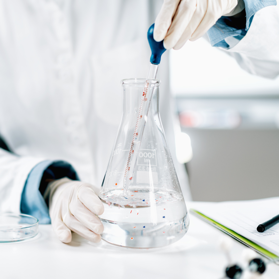 the white-lab-coated torso of a white latex gloved individual who is using a stopper to take water from a beaker. the water has visible pieces of red and blue plastic shards floating in it. 