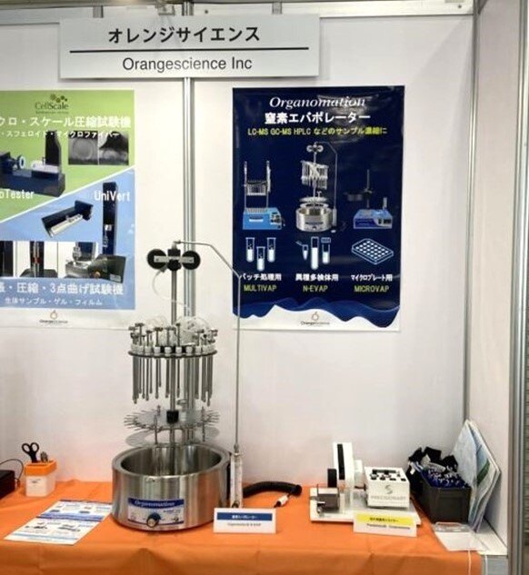 The Orange Science booth at JASIS, displaying a N-EVAP evaporator on an orange tablecloth. The table also has flyers and other small items on it, and posters hang on the wall behind the booth for various Organomation units. 