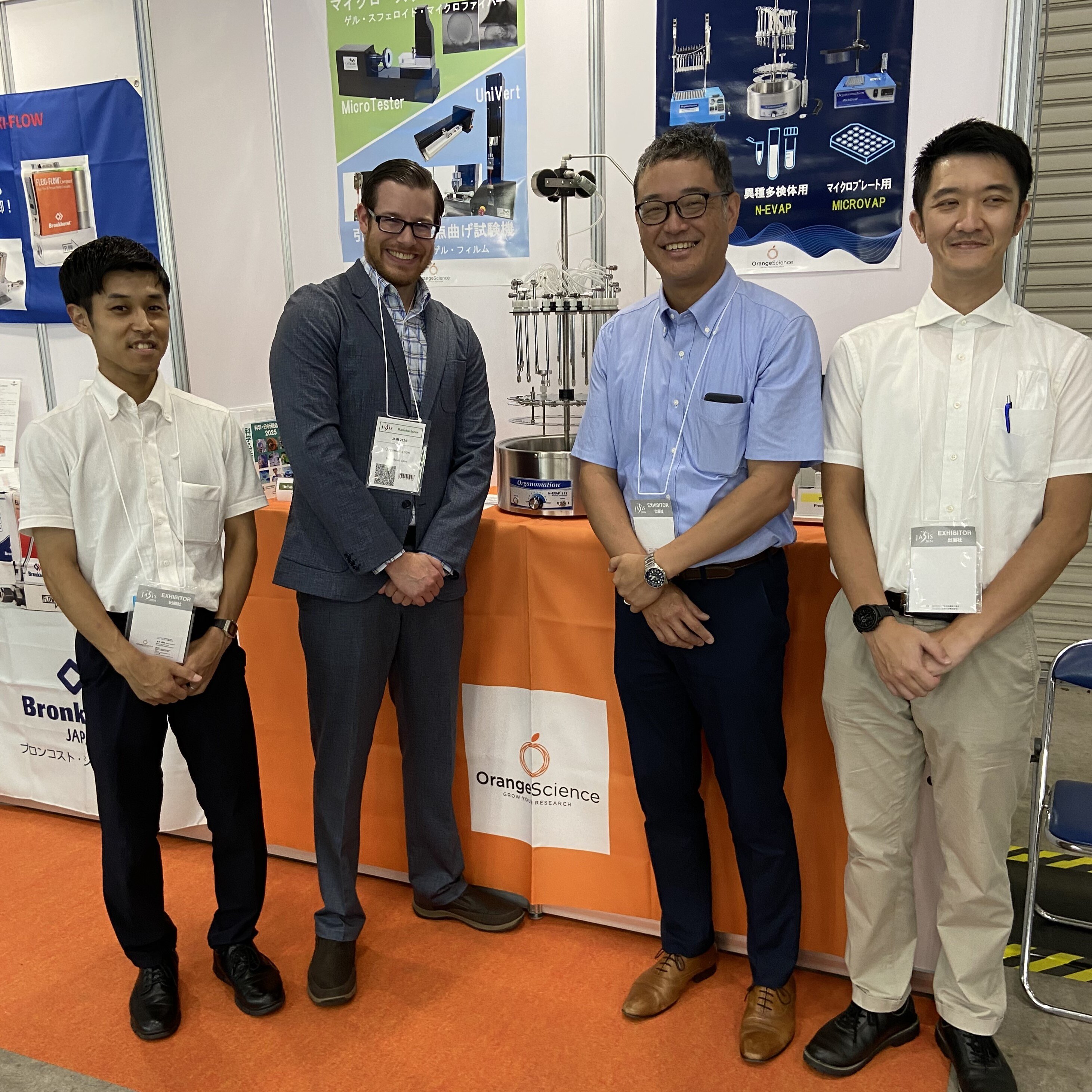 A group of four men at a trade show, standing on an orange carpet in front of a booth displaying a blowdown evaporator. 